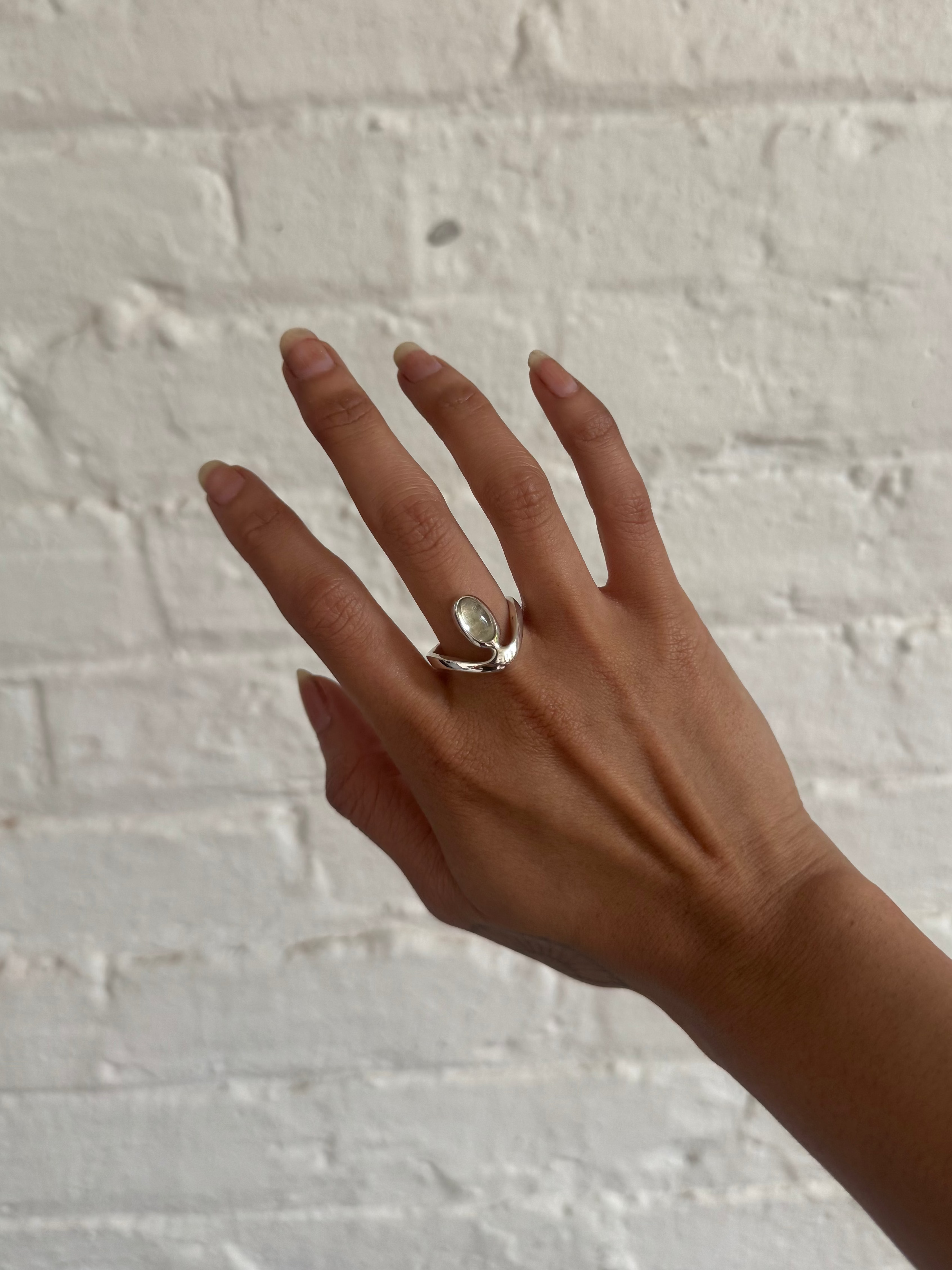 A silver ring with a clear oval gemstone worn on a person's hand, set against a white painted brick wall.
