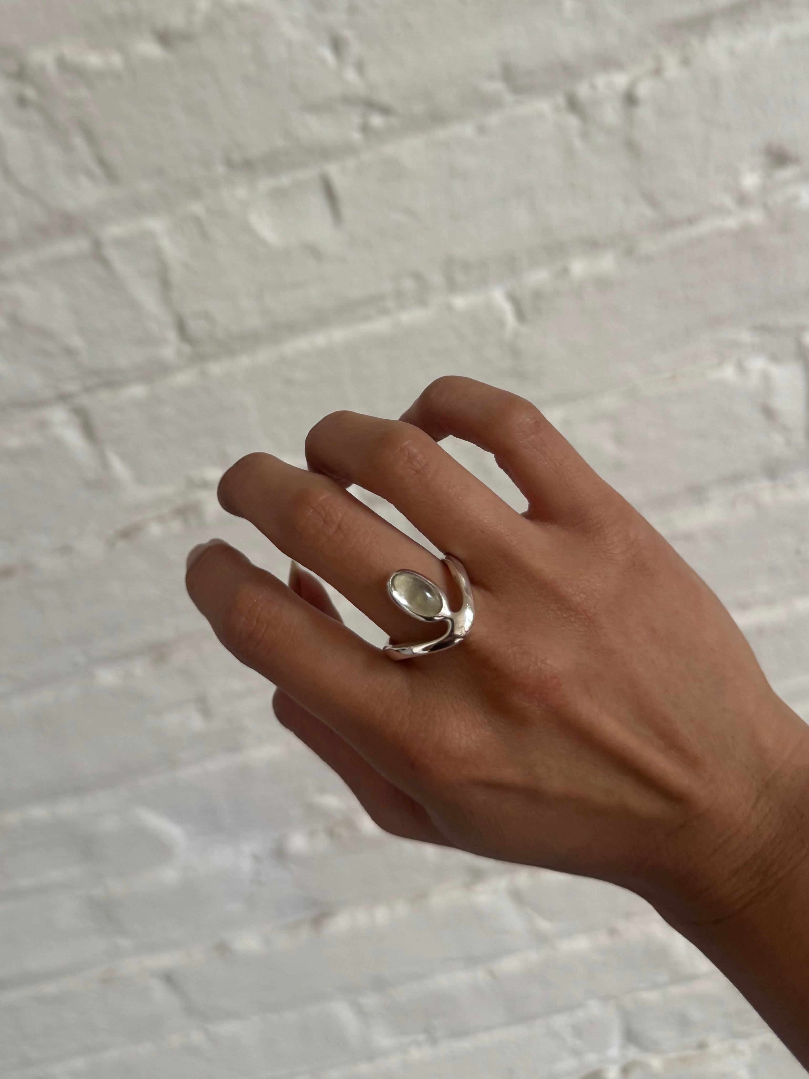A silver-toned ring with a small oval clear stone worn on a person's hand, set against a white painted brick wall.