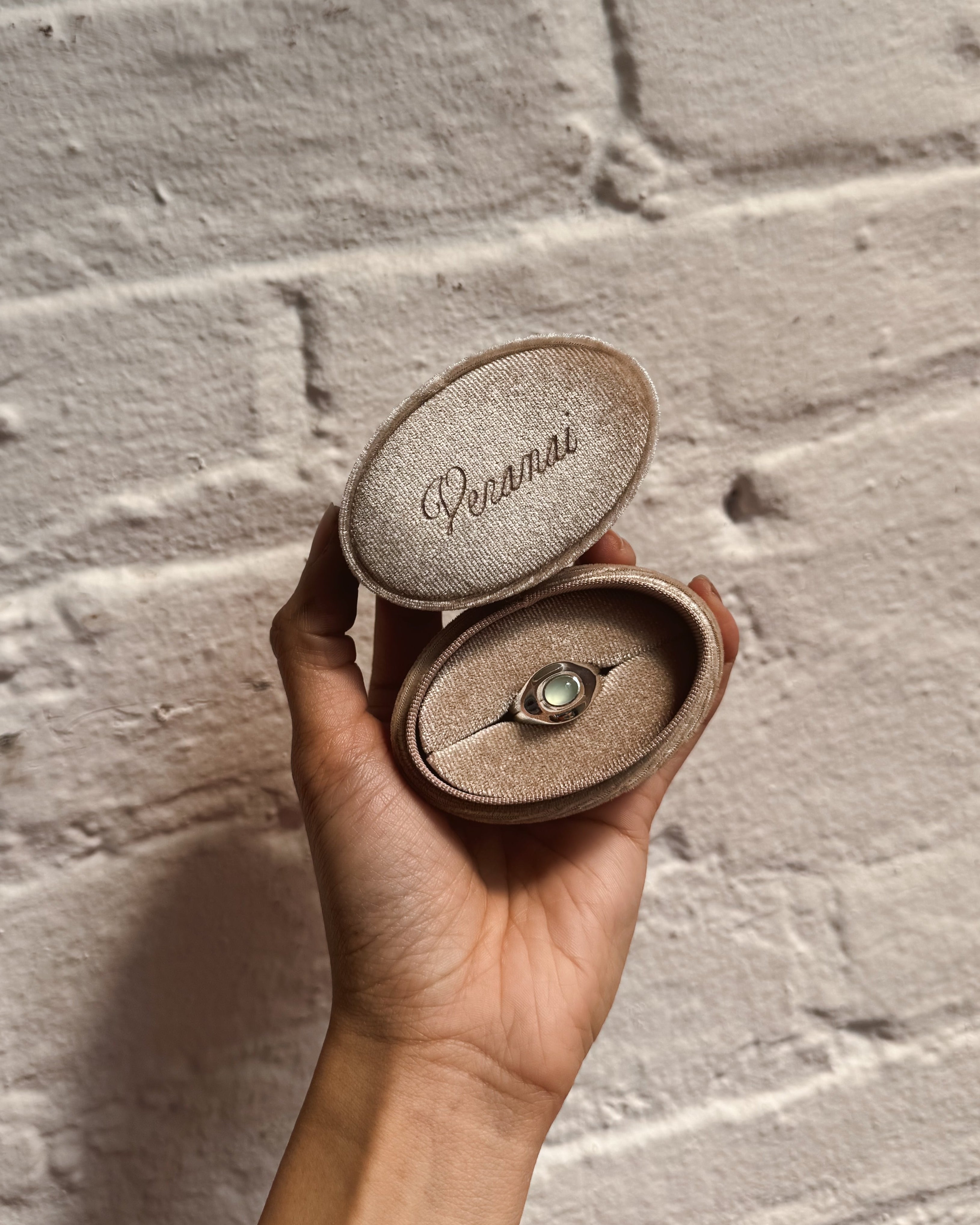 Hand holding a small, oval, peach velvet jewelry box with 'veranai' engraved on it against a textured beige wall.